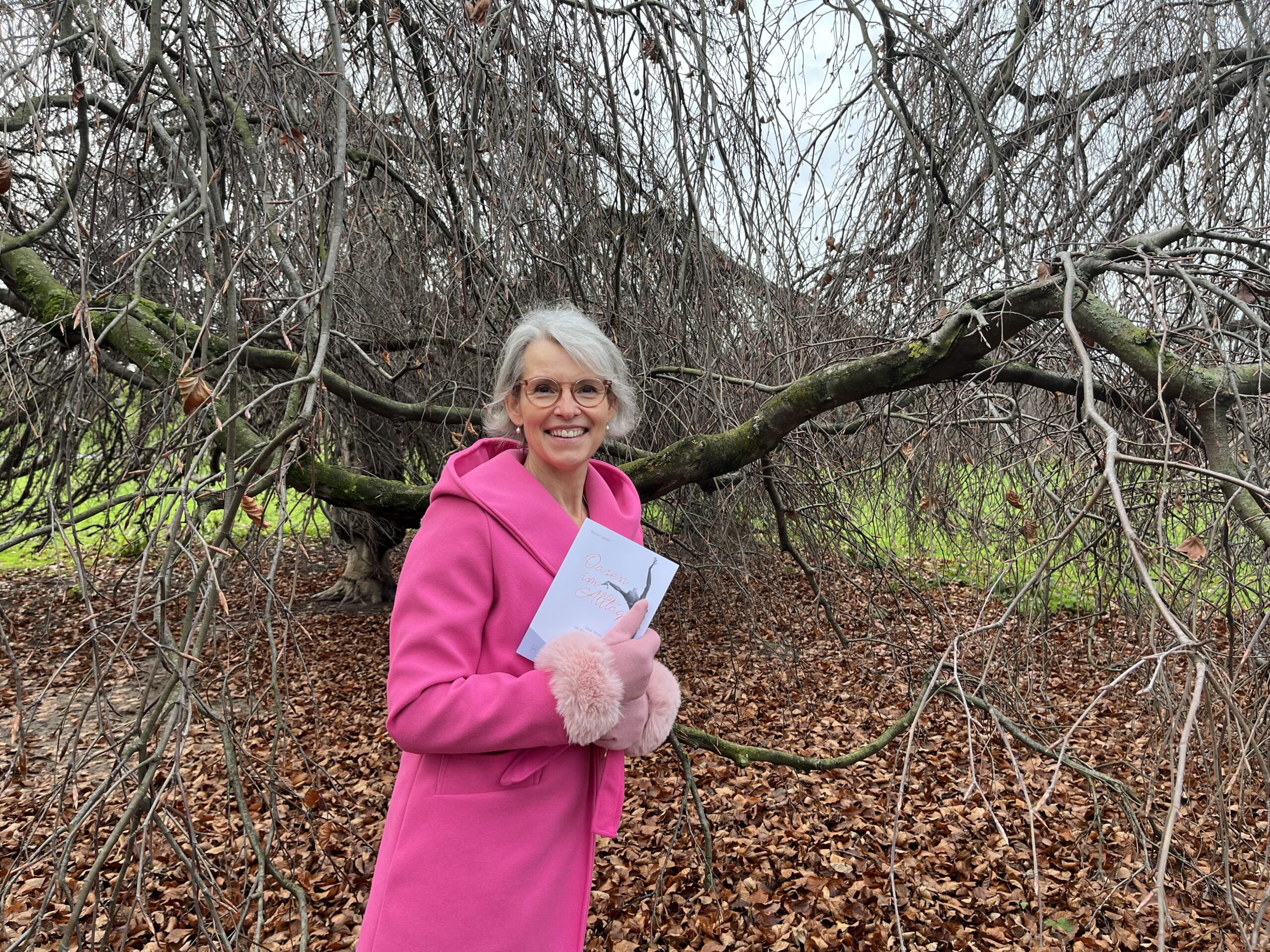 Dr. Katrien Jacobi im pinken Mantel und mit ihrem Buch vor einem winterlich blattlosen Baum