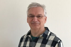 Mann mit kurzen weißen Haaren, Brille und schwarz-weiß-kariertem Flanell-Hemd lächelt in die Kamera